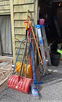 Photo showing group of yard tools including two snow shovels, rakes, pitchfork, and Werner ladder leaning against building
