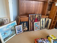 Overview of all seven art pieces arranged on the floor against a wood-paneled wall, showing various sizes and mediums including paintings and tapestry.