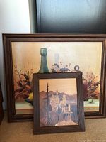 Two framed art pieces leaning against wall, showing a large still life painting behind a smaller framed wood inlay castle scene artwork.