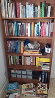 Full shelf view showing a variety of hardcover and paperback books from various categories, stacked with some boxed sets and loose papers.