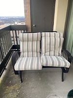 Full front view of two-seater metal glider with striped cushions on balcony, showing seating and metal armrests.