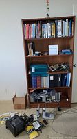 Wooden bookshelf with books, binders, phones, calculators, label printer, speakers, and cords on floor