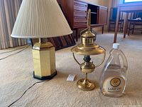 Two table lamps on carpet: a brass oil lamp with curved handle and chimney holder next to a bottle of lamp oil and gold-toned metal base lamp with white pleated shade