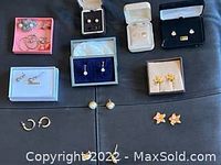 Assorted sets of earrings laid out on a flat surface including small hoops, stud earrings in various shapes such as stars and flowers, and pearl drop earrings, some in small boxes.