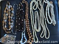 Photo showing seven assorted necklaces laid out on a black surface: a multi-colored beige bead necklace with irregular shapes, a dark brown and pink glass bead necklace, a natural wooden bead necklace, two white faux pearl necklaces of different lengths, a beige-toned pearl necklace, and a long clear beaded necklace.