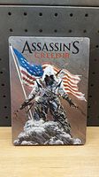 Front view of Assassin's Creed III steelbook for PS3 showing American flag and character Connor Kenway with game title.