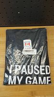Front of boxed black t-shirt showing part of white text "I PAUSED MY GAME"