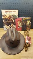 Full lot showing all items: three Indiana Jones books, brown toy fedora hat, and toy whip with tag on a wooden table with black pegboard background.