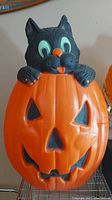 Front view showing black cat poking head and paws out of the orange jack-o-lantern pumpkin with carved triangle eyes and mouth.