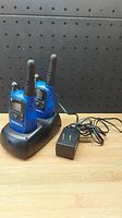 Pair of blue Motorola walkie talkies seated in black dual charging dock next to AC power adapter on wooden surface with black pegboard backdrop.