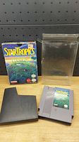 Overview photo showing Nintendo Startropics game cartridge, original box, black plastic sleeve, and clear protective box cover