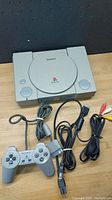 Top view of original grey Sony PlayStation console with PlayStation logo, cable and AV cables coiled on table.