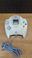 White Sega Dreamcast wired controller with analog stick, D-pad, four colored buttons, and cord on wooden surface.