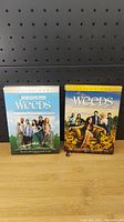 Front view of Weeds Season 1 and Season 2 DVD box sets placed side by side on a wooden surface against a pegboard background.