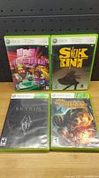 Front of four Xbox 360 game cases showing cover art for Skyrim, Big Bumpin', Sneak King, and Cabela's Dangerous Hunts 2011.