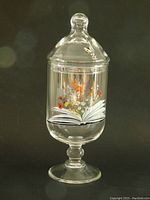 Front view of clear glass apothecary jar with floral transfers rising from an open book and a bumble bee on the removable lid against a black background.