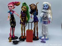 Four Monster High dolls standing with two accessories centered: a red book with dragon design and a small broom. All dolls have stands.