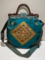 Front view of the green fabric bag with central golden yellow embroidered diamond pattern, green fabric shoulder strap, and brown wicker purse handles.