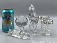 Six assorted large faceted glass or crystal bottle stoppers shown with soda can for size reference. Varied shapes include pointed, spherical, oval and elongated geometric faceting.