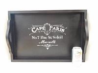 Front view of rectangular black wooden serving tray with built-in handles, featuring cream vintage stencil reading 'Cafe Paris No. 7 Rue St Soleil Marseille' with filigree embellishments. Includes a can for size reference.