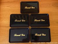 Photo of the set of five black plastic trays with gold "Thank You" lettering, arranged flat on wooden surface showing top view of each tray.