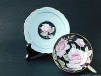 Photo showing front view of saucer on stand and teacup interior with three pink cabbage roses on black background, gold edging visible