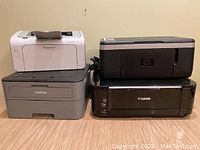 Four printers on wood floor against beige wall, stacked two by two: white HP LaserJet P1005 and gray Brother HL-L2320D on left; black HP Deskjet F4180 atop black Canon PIXMA MG5220 on right.