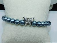 Black pearl bracelet displayed on a cushion, showing the single strand of black pearls and the silver-tone puff butterfly charm central on the strand.