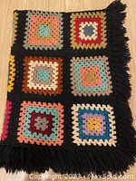 Close-up photo of folded multi-coloured crocheted granny squares throw blanket on wooden floor, showing detail of squares and black fringe.