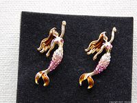 Pair of whimsical mermaid stud earrings on a black felt background showing details of gold-tone metal, enamel hair and tailfin, and sparkling rhinestone bodies.
