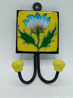 Front view of the hand-painted ceramic tile coat hook showing the blue and green floral design on yellow, metal frame, and dual hooks with yellow ceramic caps.