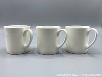 Front view of three small white bone china mugs by Wedgwood with curved handles.