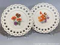 Front view of two vintage ceramic plates with hand-painted floral designs and lace-like pierced edges on display stands.