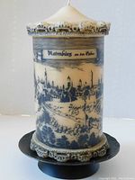 Front view of vintage pillar candle with detailed black illustration of Rothenburg ob der Tauber, showing crenulated top and bottom edges with creamy beige wax and black accents. Candle is mounted on black metal pedestal stand with scalloped rim.