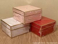 Three gift boxes stacked: white, pink, and red with gold trim, all wrapped in plastic.