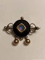 Front of antique round gold plated brooch with black onyx center and central glass or opalescent stone, gold plated scroll accents on sides, and three hanging beads.
