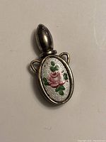 Front view showing guilloche white enamel with pink rose and green leaves design on oval bottle attached to metal stopper