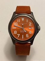 Front view of Momentum watch showing orange dial, white numerals, date window at 3 o'clock, and silver hands.