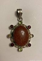 Front view of the sterling silver pendant showing the large oval goldstone central cabochon and eight surrounding round garnet and peridot accent stones.