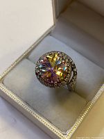 Top and angle view of sterling silver ring with large rainbow multifaceted gemstone and smaller clear stones in a white ring box.