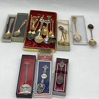 Wide view of 22 assorted souvenir spoons in sets and boxes, mostly gold plated with enamel highlights, laid out showing varied designs and sizes.