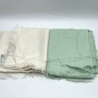 Two folded blanket scarves, one ivory and one mint green, placed side by side showing their colors and fringed edges.