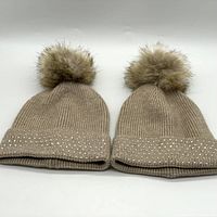 Two oatmeal knit beanies with crystal embellishments on cuffs and faux fur pom poms, shown side by side.