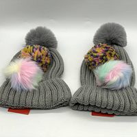 Two grey BP knitted beanies shown side by side, each with one grey pom on top and two interchangeable poms: a multi-colored yarn pom and a pastel-colored faux fur pom