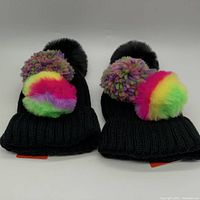 Two black knitted BP beanies shown side by side each with a large black pom on top and an additional colorful fuzzy pom attached.