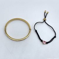 Two Kate Spade bracelets: gold-tone bangle and black cord slider with pink heart charm.