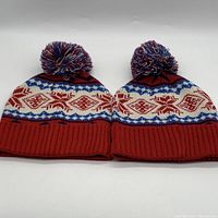 Two red Fair Isle patterned toques with multicolored pom-poms displayed side-by-side showing full front view.