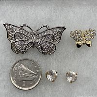 All four items shown together with a Canadian coin for scale: large silver butterfly pin, small gold butterfly pin, and 14K gold stud earrings with heart-shaped Swarovski crystals