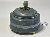 Side angled view of the antique aluminum powder puff style music box showing textured surface, horizontal banding, brass feet and green finial knob on top.
