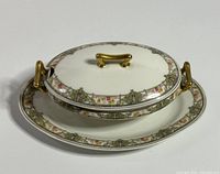 Closed covered serving dish on matching oval platter, side view showing gold-tone handles and ornate floral rims